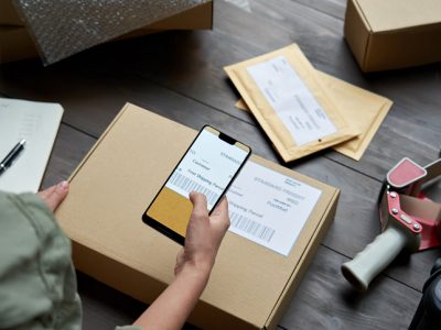 Female warehouse worker, seller, dropshipping small business owner holding phone scanning retail package postal parcel barcode on ecommerce shipping box label on smartphone using mobile app, close up.